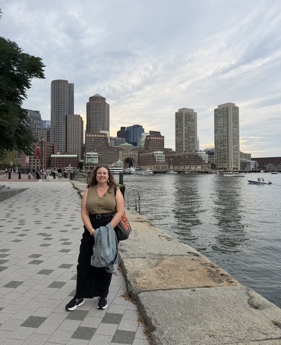 Miriam smiling on the Boston Harborwalk with the skyline behind her